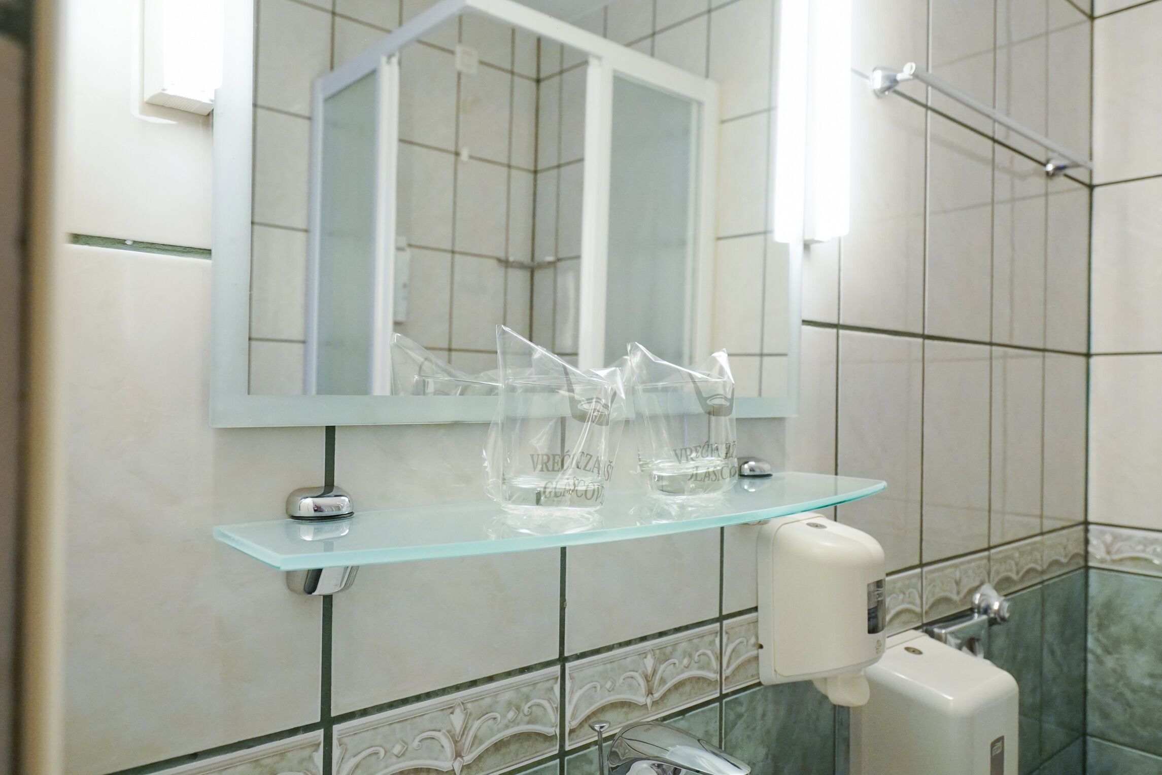 Une salle de bain avec une étagère en verre contenant des bouteilles transparentes, un miroir et un distributeur de savon.