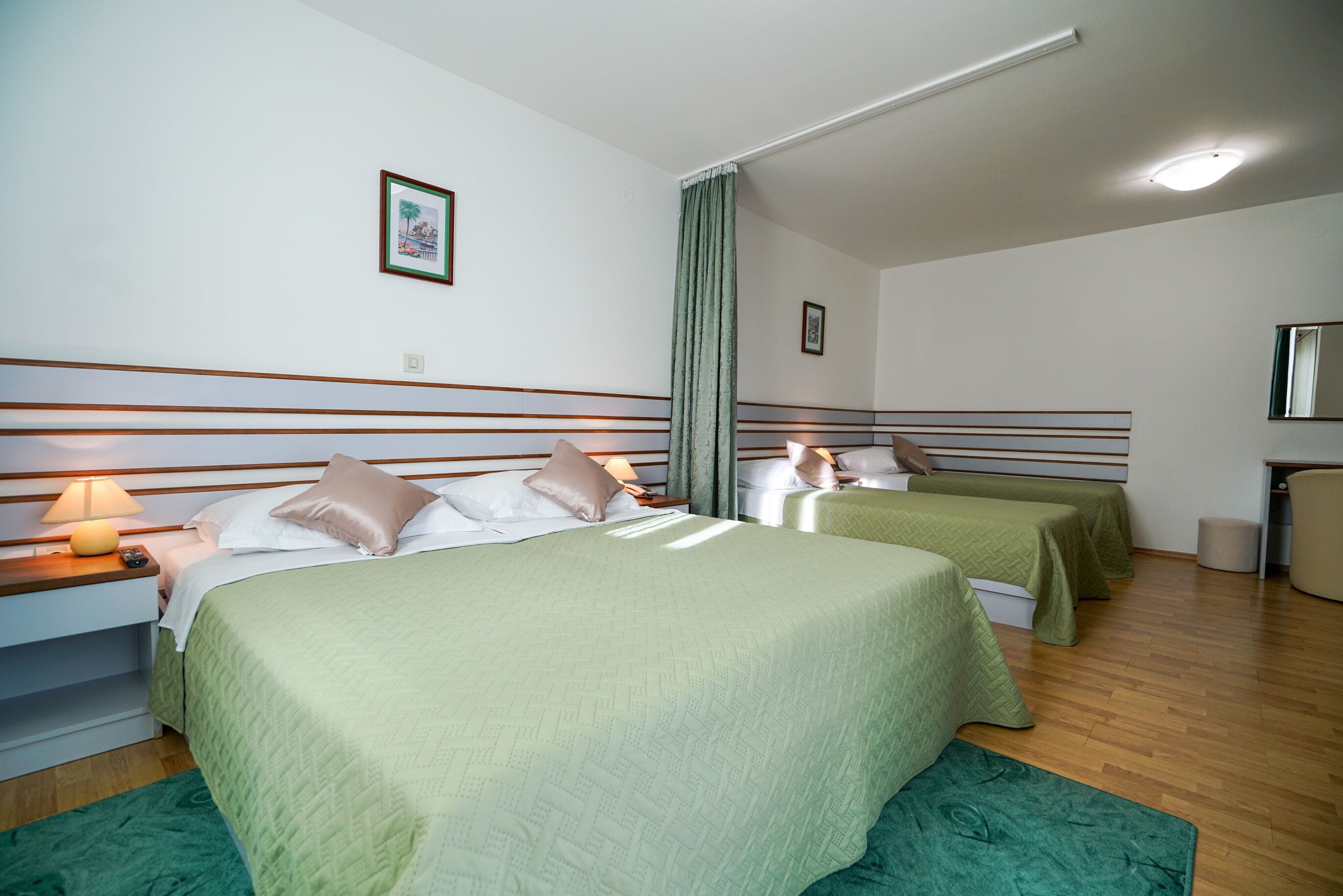 Camera d'albergo con due letti, tappeto verde, pavimento in legno e comodino con lampada.
