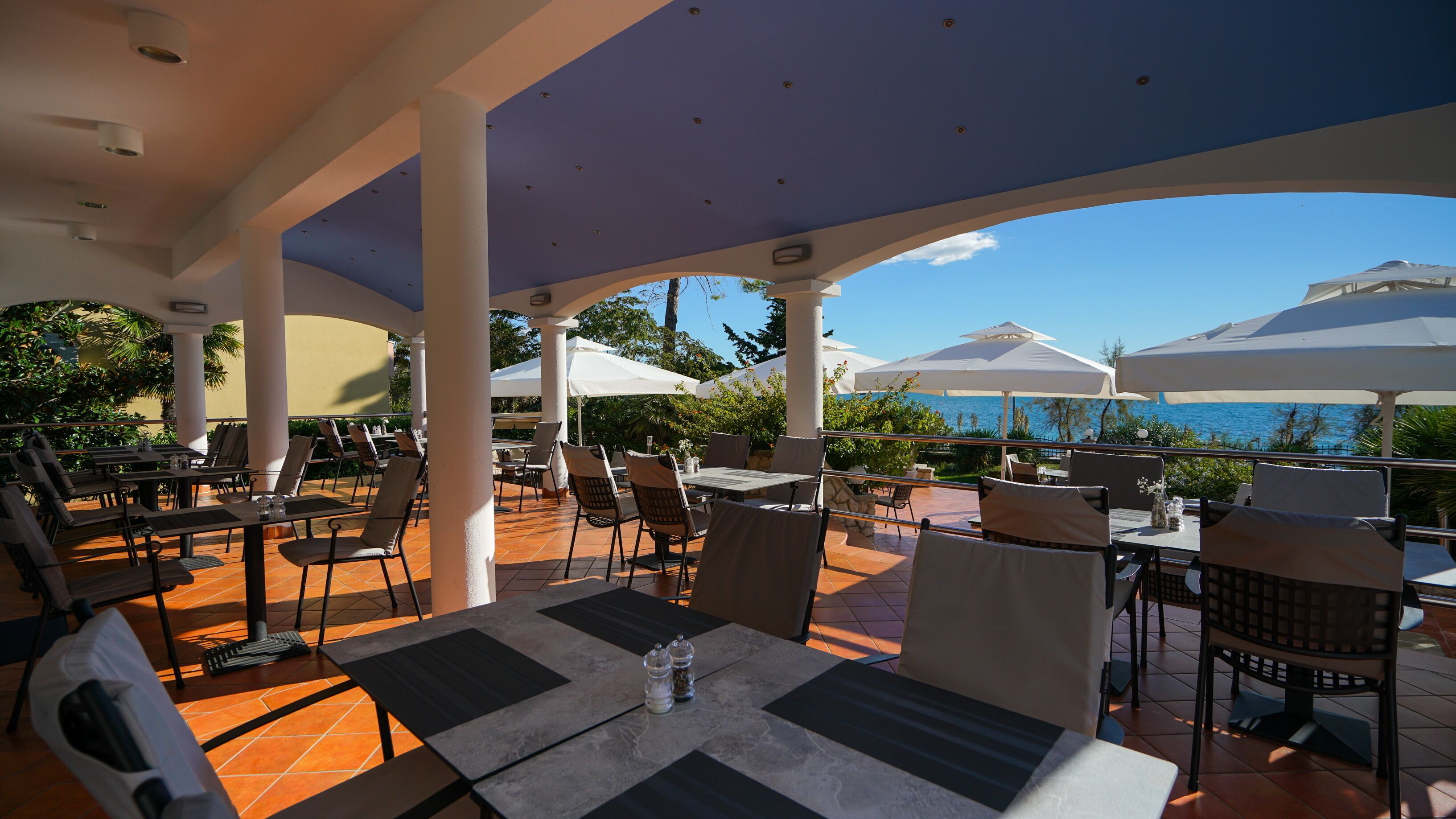 An outdoor patio with tables and chairs, umbrellas, and a view of the sea.