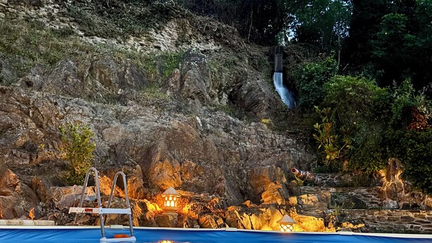 Ein Schwimmbecken mit felsigem Hintergrund, einem Wasserfall und beleuchteten Bauwerken.