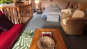 Ein gemütliches Schlafzimmer im Dachgeschoss mit Holzbalkendecke, einem Bett mit gestreifter Bettwäsche, einem Korbsessel und einem roten Sofa.