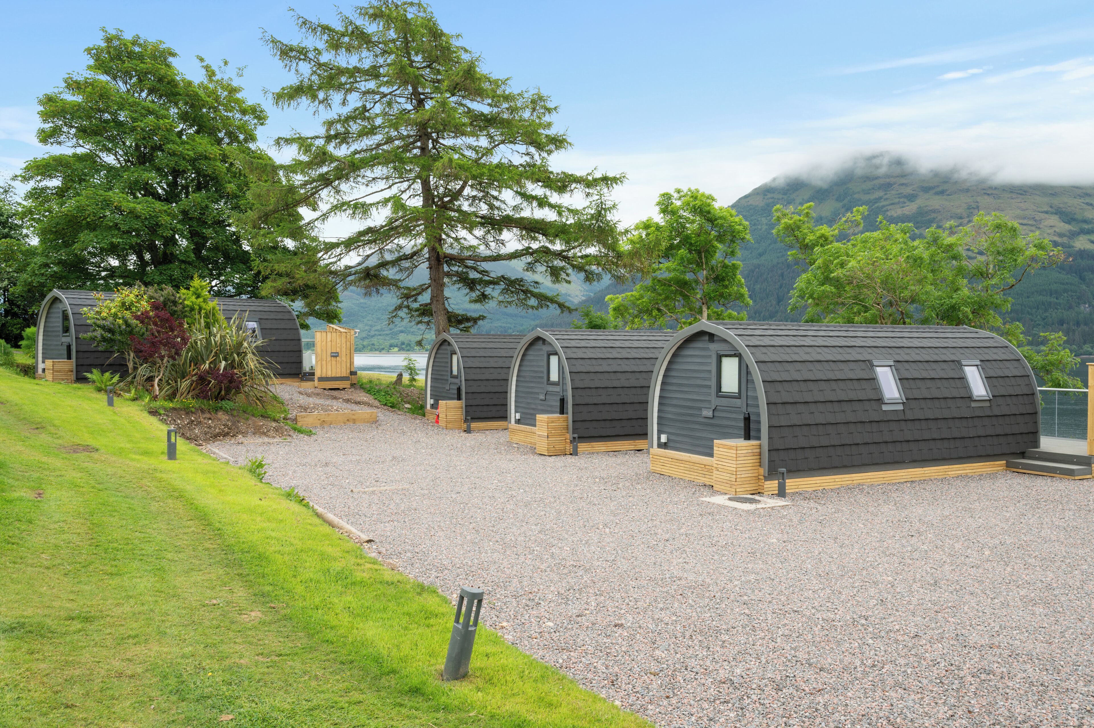 A row of modern, curved-roofed cabins with wooden decks, set against a backdrop of lush greenery and a mountain.