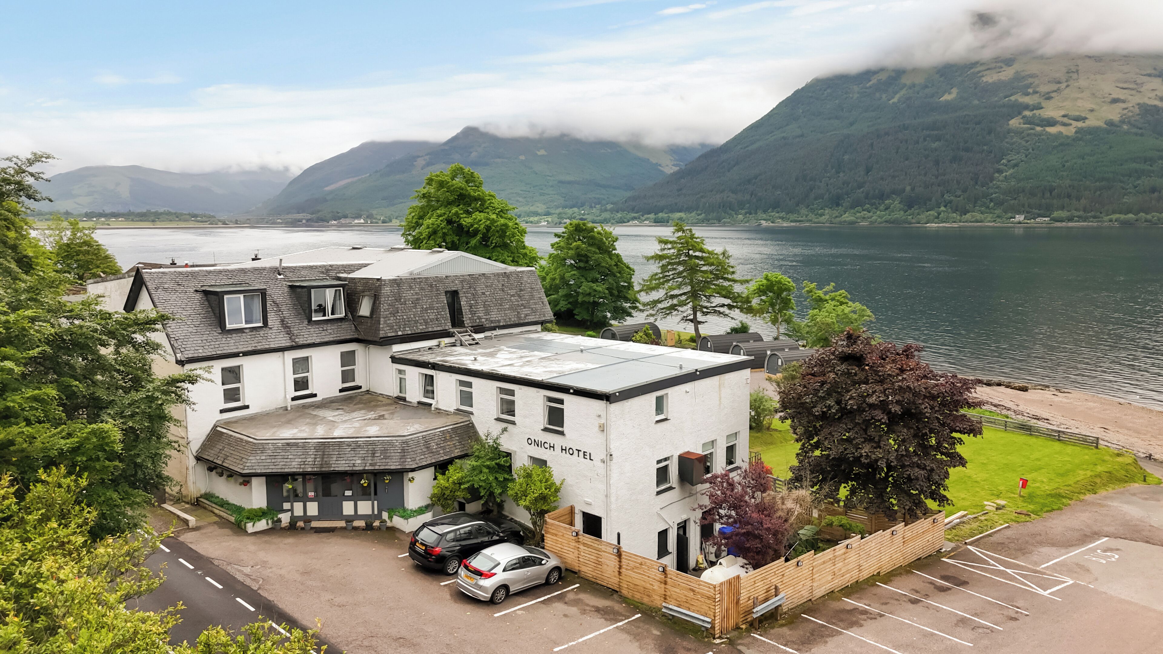 A hotel with a white facade, a parking area, and a view of a lake and mountains.