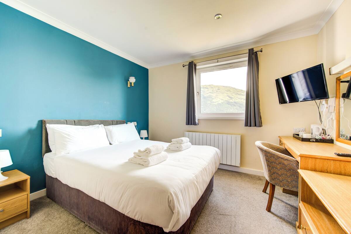 Una habitación de hotel con cama, escritorio, silla, televisor y ventana con cortinas.