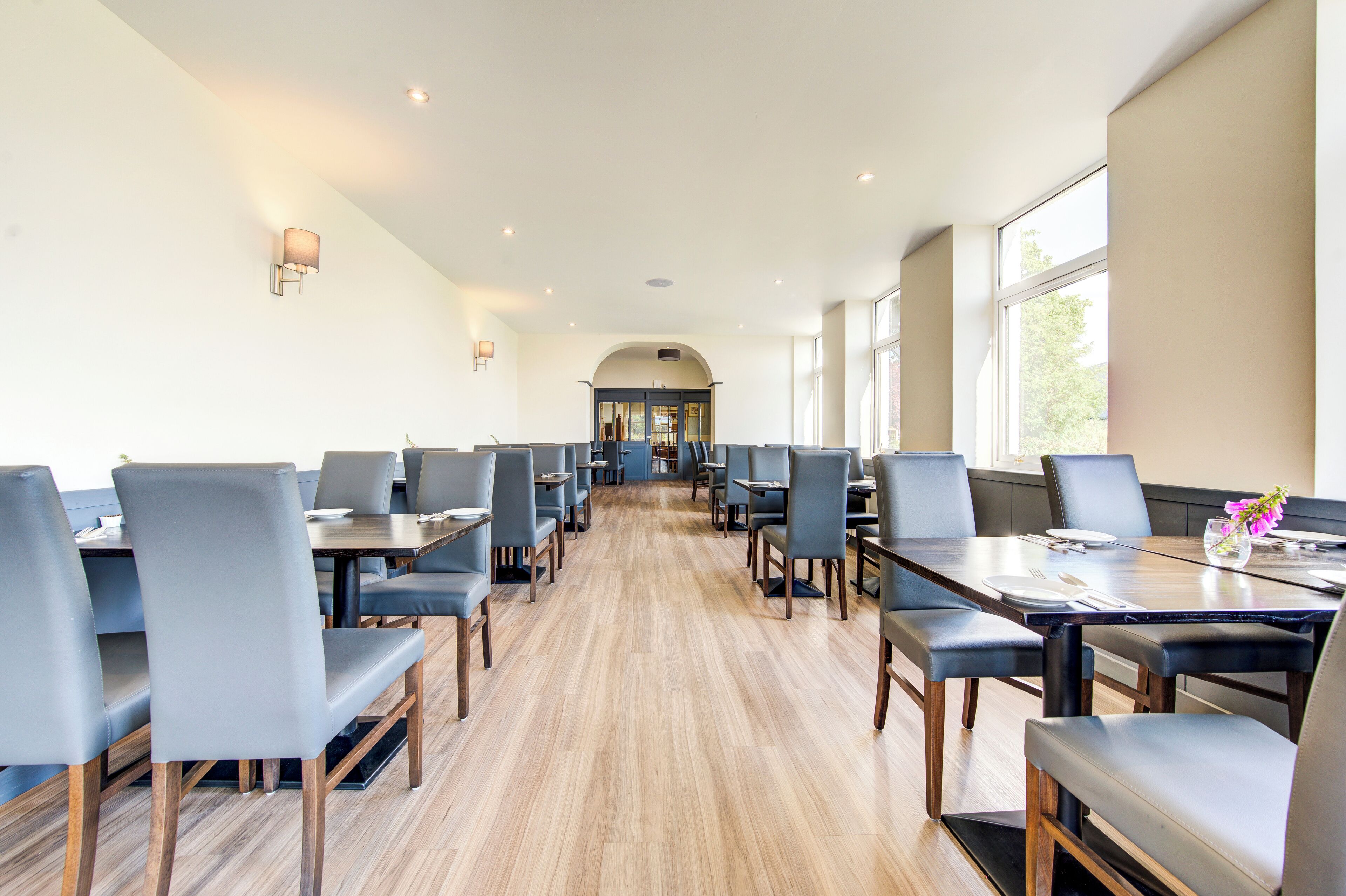 A spacious dining area with long tables and chairs, a wooden floor, and large windows allowing natural light.