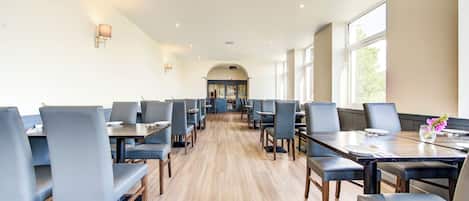 A spacious dining area with long tables and chairs, a wooden floor, and large windows allowing natural light.