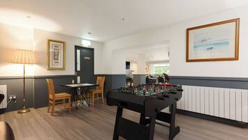 A room with a foosball table, a lamp, framed pictures, and a view of the kitchen.