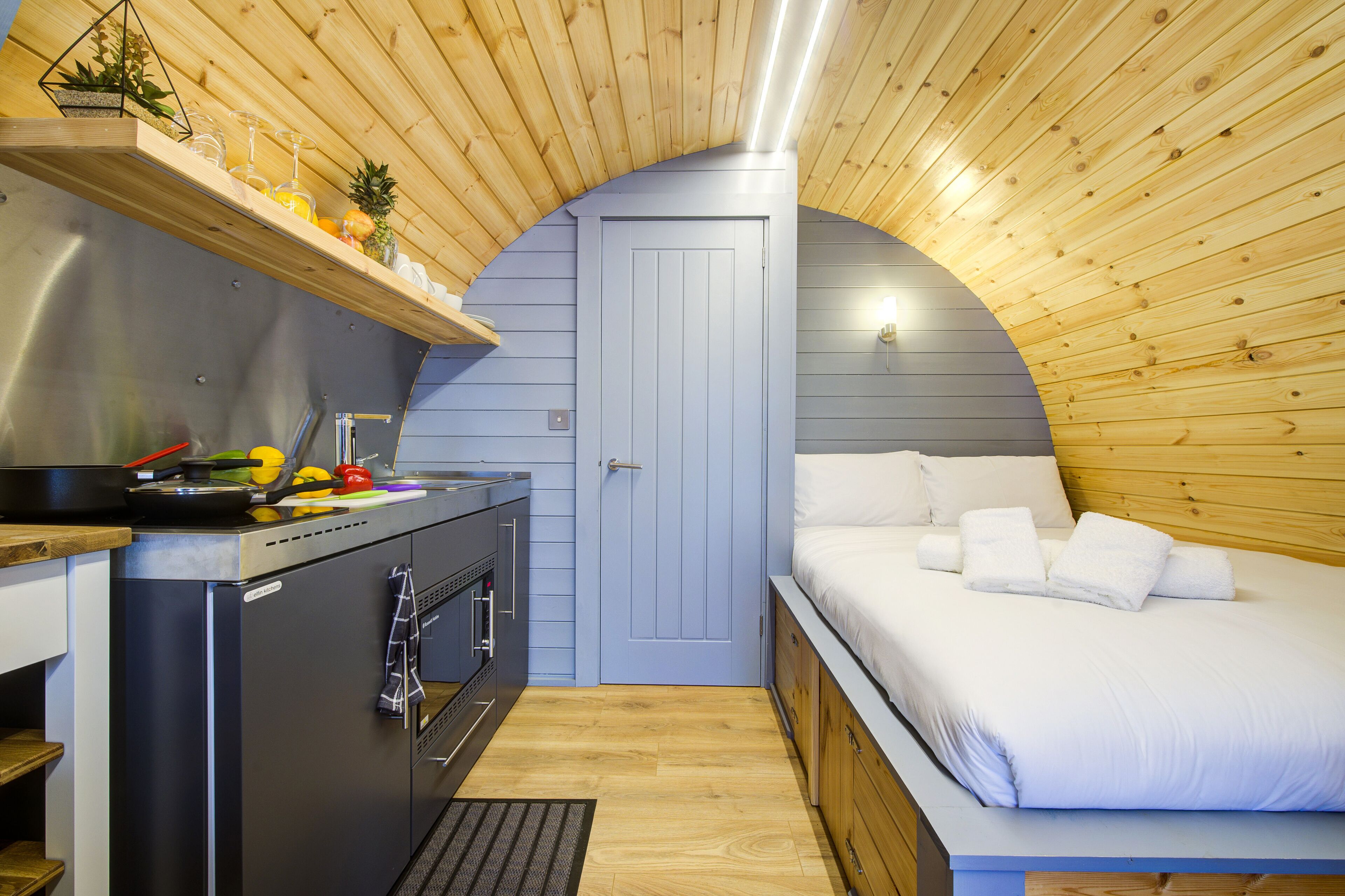Een compacte kamer met een kitchenette, een bed en een gebogen houten plafond.