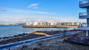 A coastal view with modern buildings, a sandy beach, and a rocky shoreline. - Seaside Paradise Apartment (Garðabær)