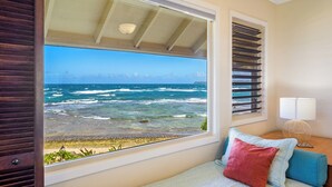 Un dormitorio con un ventanal grande con vista al océano, una cama con almohadas, una mesita de noche con una lámpara y vistas a la playa y al horizonte de la ciudad.