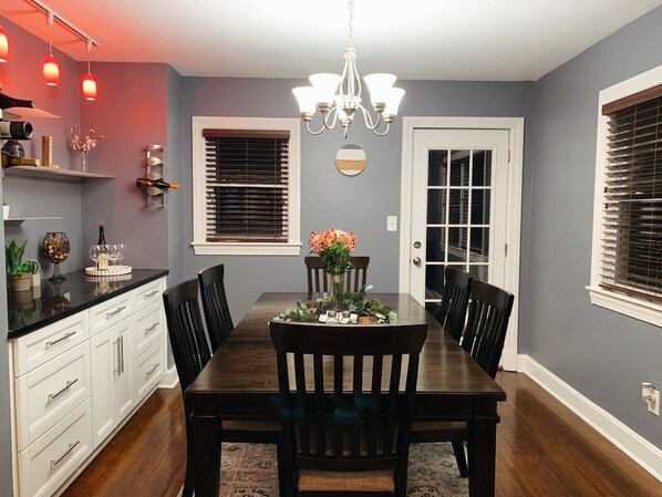 Une salle à manger avec une table en bois, des chaises et un lustre. On y trouve des fenêtres équipées de stores, une porte et une étagère ornée d’objets décoratifs.