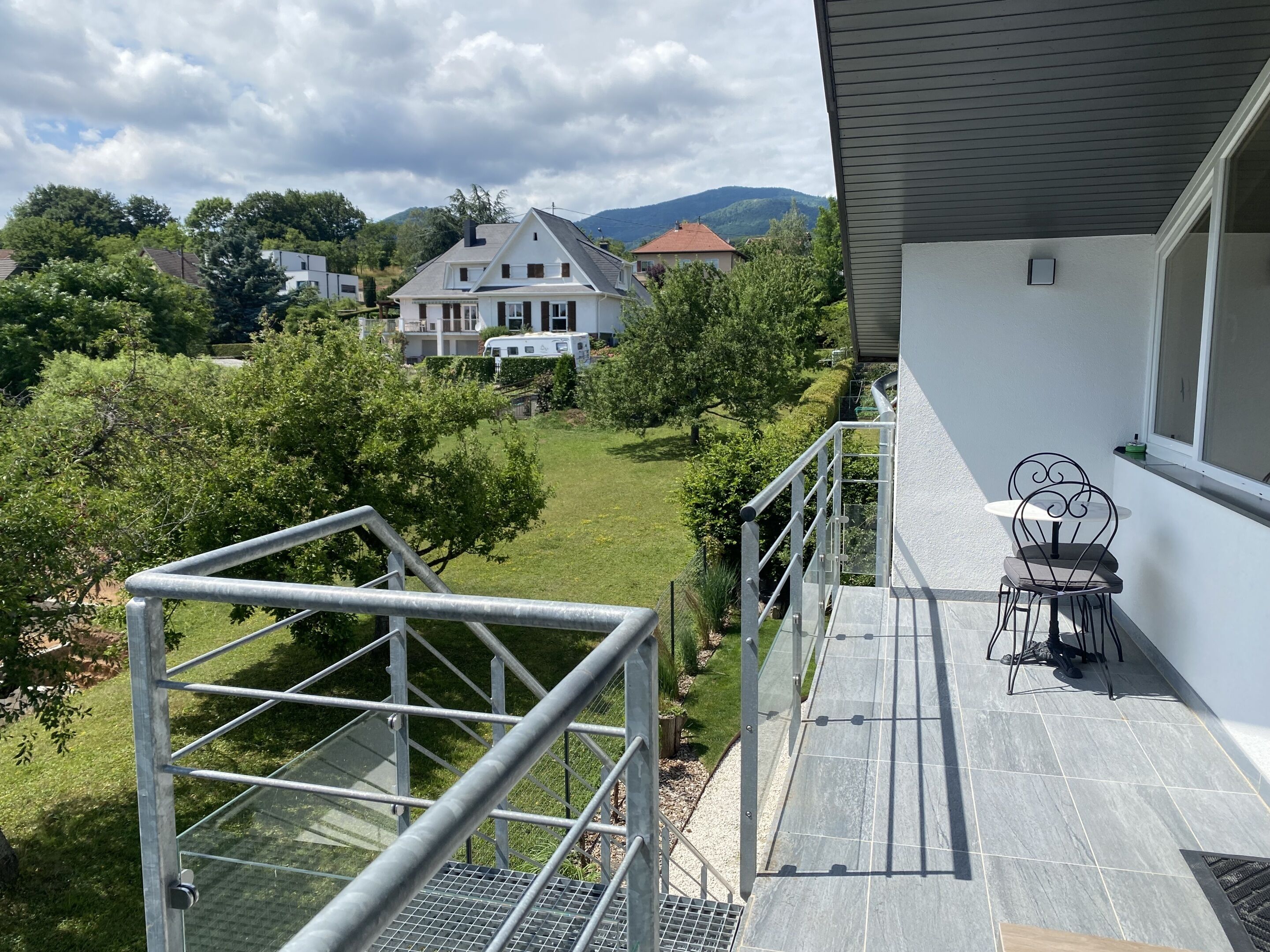 Uma varanda com guarda-corpo de metal, uma mesa pequena com uma cadeira e vista para uma casa e vegetação ao fundo.