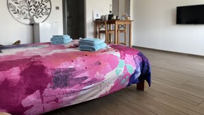 Une chambre avec une couvre-lit aux couleurs vives, un plancher en bois, un bureau avec une chaise et un téléviseur à écran plat fixé au mur.