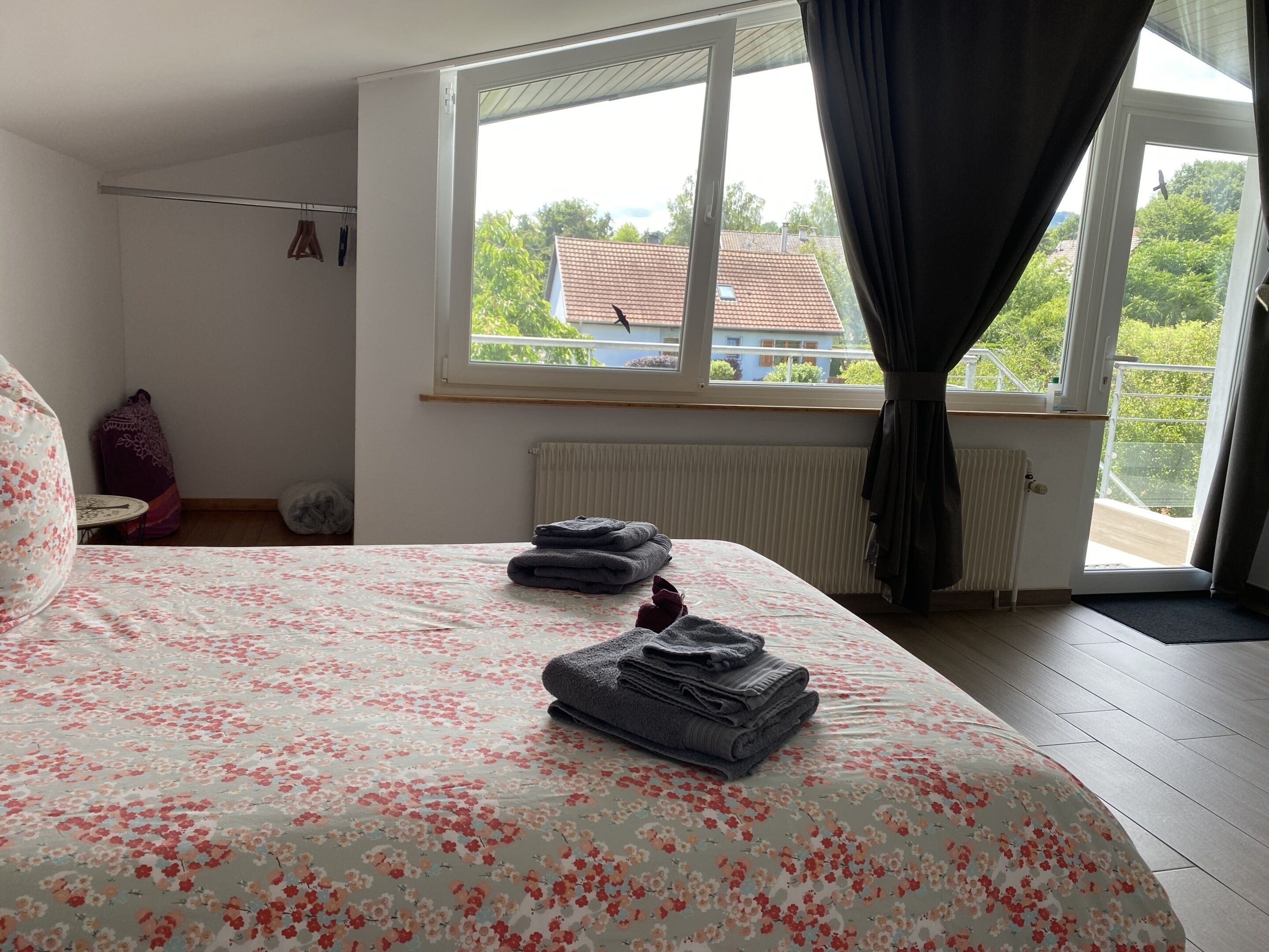 A bedroom with a bed, a window with curtains, a radiator, and a view of a house and trees outside.