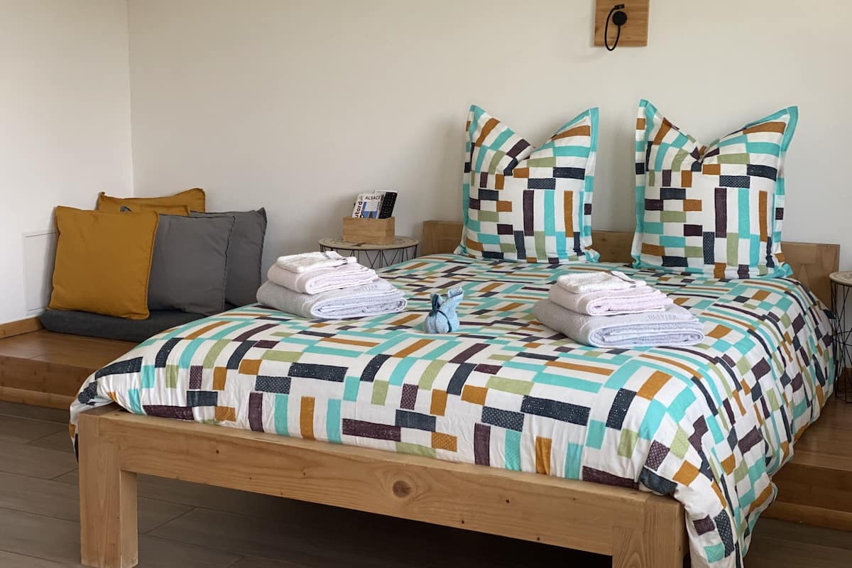 A neatly made bed with a colorful quilt, a wooden bench, and a small table with a box of toiletries.