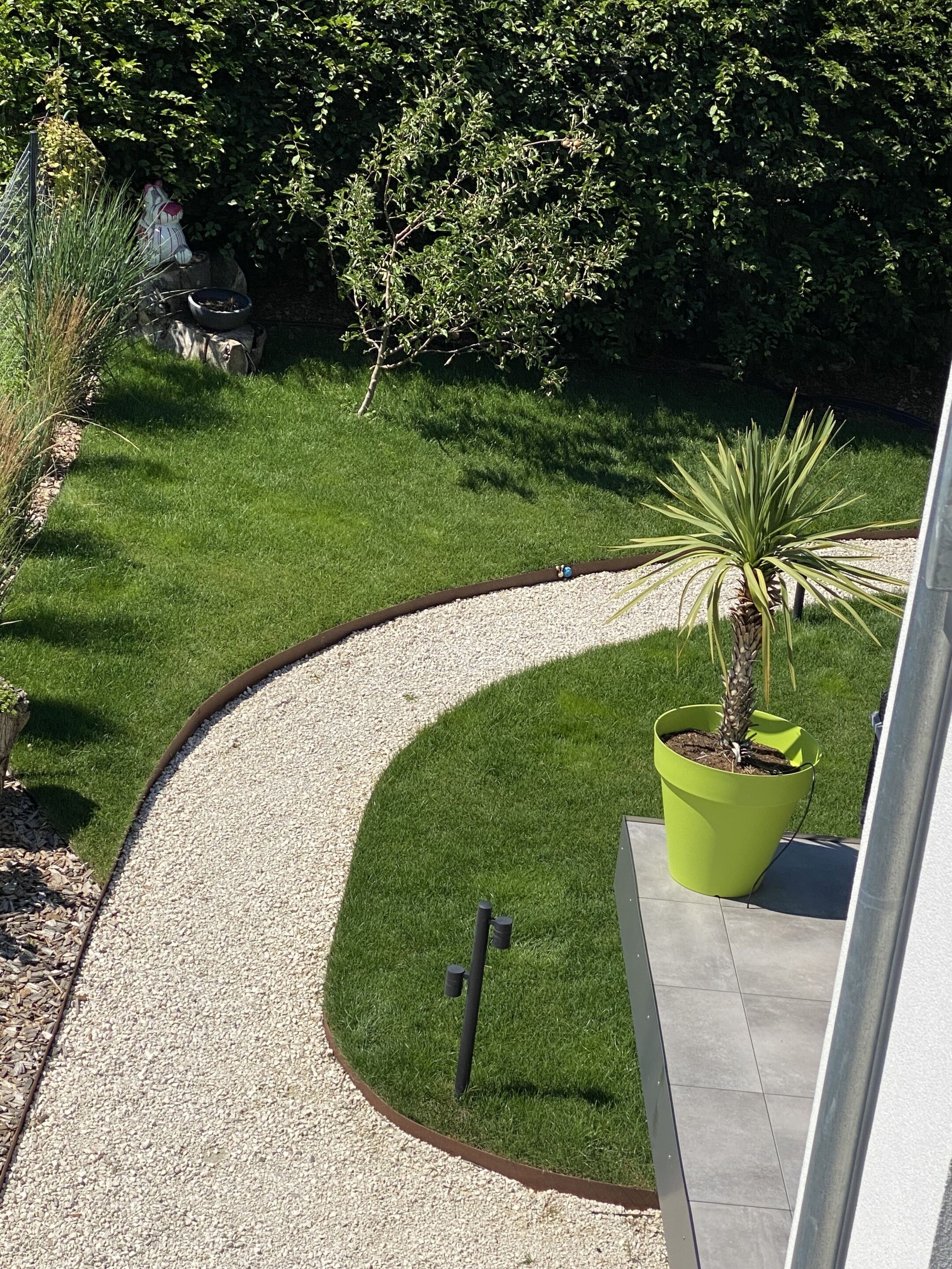 A landscaped garden with a curved gravel path, a large green planter with a palm tree, and a small statue of a dog.