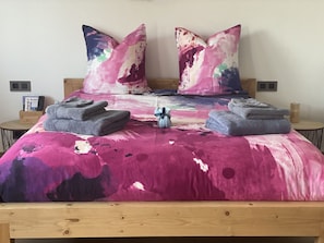 Ein Bett mit bunter Bettdecke, zwei Kissen und ein kleiner Nachttisch mit Fernbedienung und Kerze.