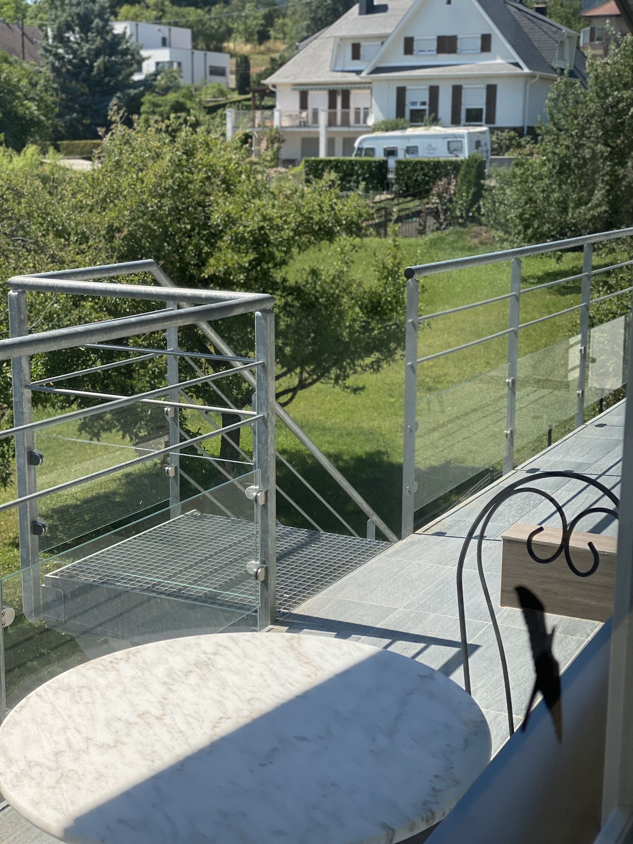 Un balcon doté d’une balustrade en métal, une table en marbre blanc et une vue sur un quartier résidentiel avec des maisons et de la verdure.