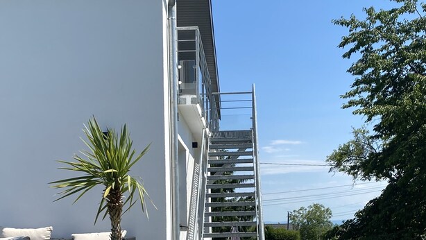 Une maison moderne dotée d’un patio couvert, d’une plante verte en pot, d’un escalier en métal et d’un chemin de gravier.