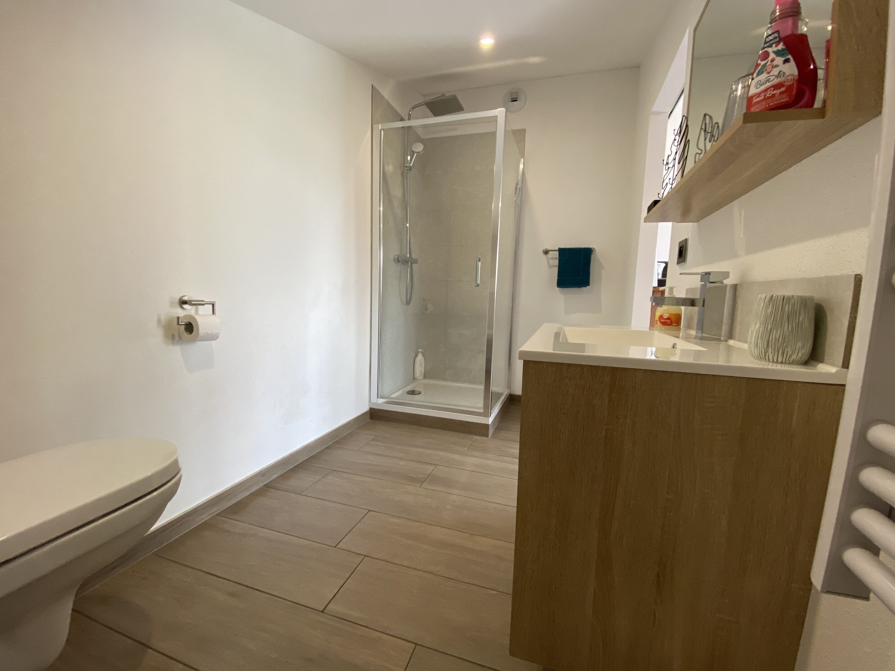 A modern bathroom with a toilet, a glass-enclosed shower, a wooden vanity, and a shelf with toiletries.