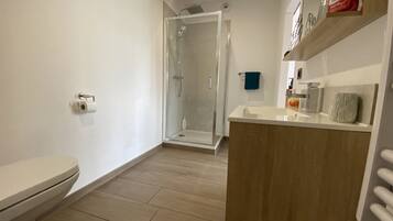 A modern bathroom with a toilet, a glass-enclosed shower, a wooden vanity, and a shelf with toiletries.