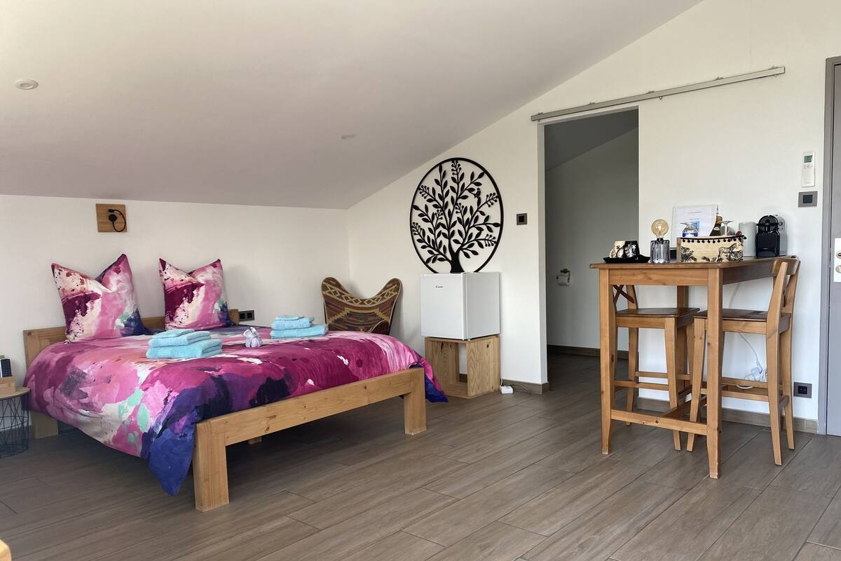 A bedroom with a bed, wooden headboard, a wall-mounted tree decoration, a small table with a kettle, and a door leading to another room.