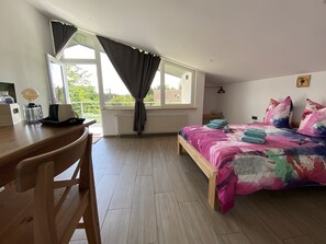 Un dormitorio con cama, escritorio y silla. Hay una ventana con cortinas, un radiador y vistas al exterior con vegetación.