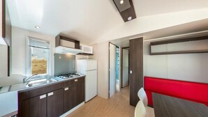 A compact kitchen with dark wood cabinets, a gas stove, and a microwave. There is a red bench and a white chair in the dining area. - Camping le Chenal (Saint-Jean-de-Monts)