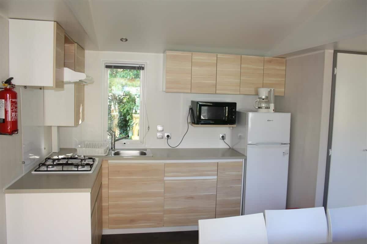 Una cocina compacta con estufa a gas, microondas y refrigerador, armarios de madera y una ventana con vista a la vegetación.