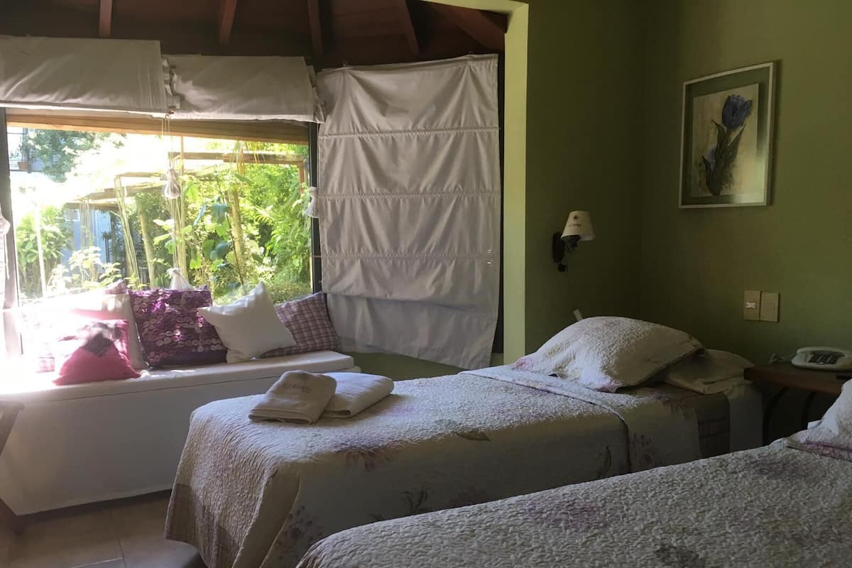 A room with two beds, a window with curtains, a wall-mounted lamp, and a painting.