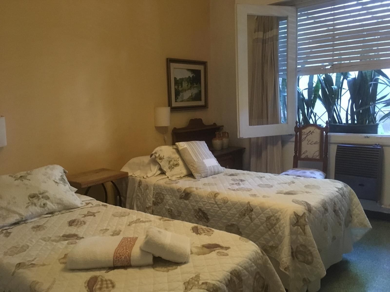 A room with two beds, a bedside table, a lamp, a framed picture on the wall, and a window with curtains.