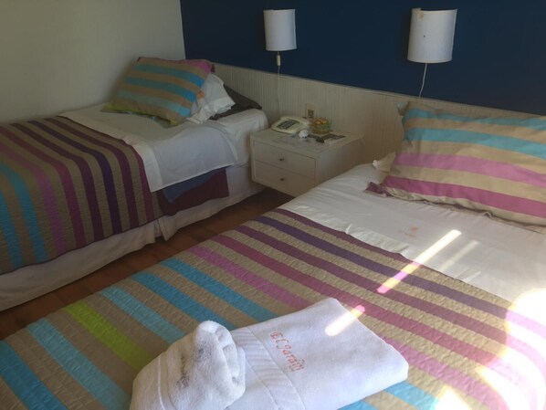 A hotel room with two beds, each with striped bedding and pillows, a bedside table with a phone and a lamp, and a nightstand with a lamp. - Hotel El Jardin (Paysandu)
