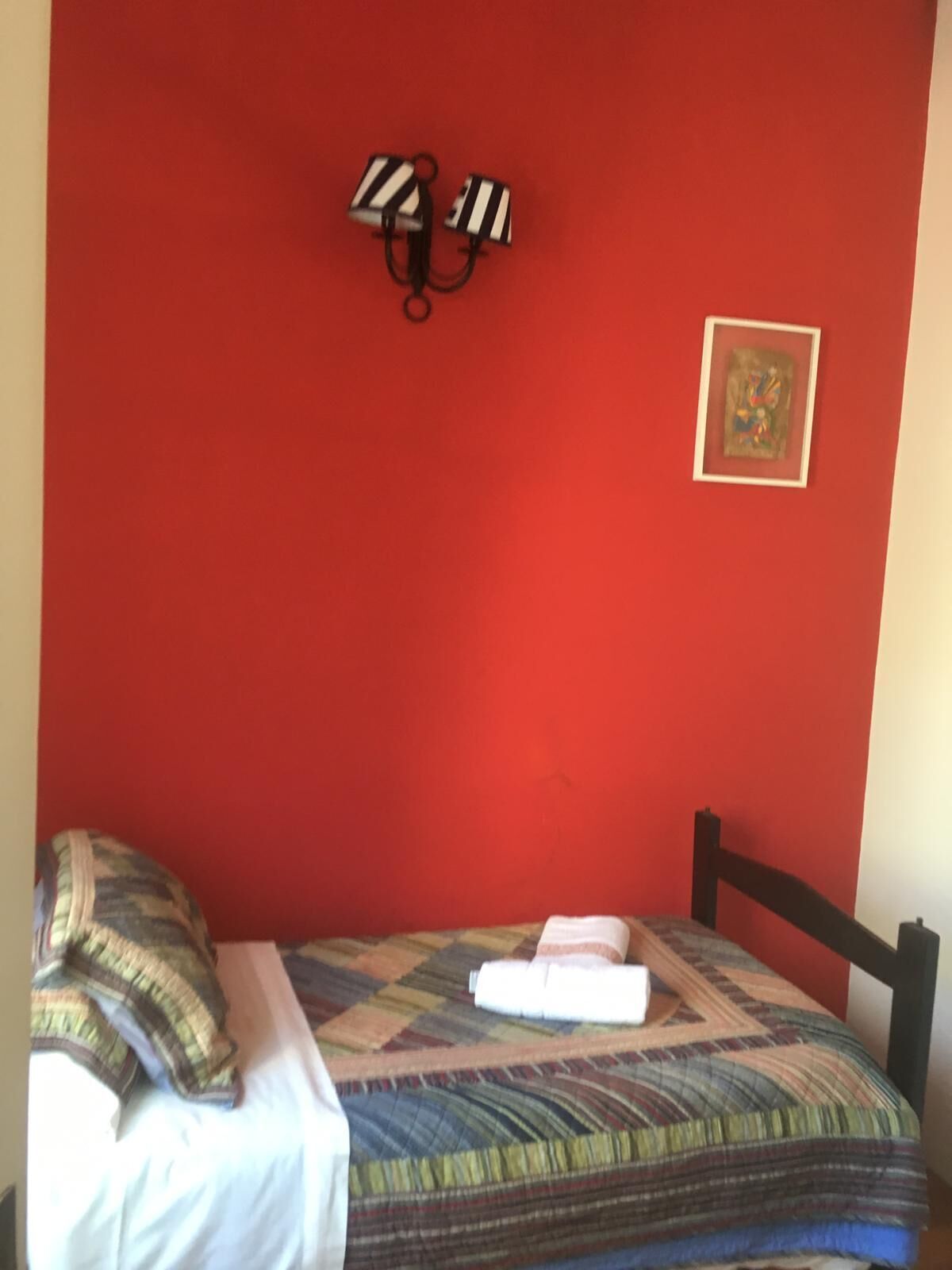 A room with a red wall, a bed with a striped pillow, a quilt, and a wall-mounted light fixture.