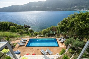Et rektangulært pool med træterrasse, omgivet af liggestole og udsigt til et stort vandområde og bjerge.