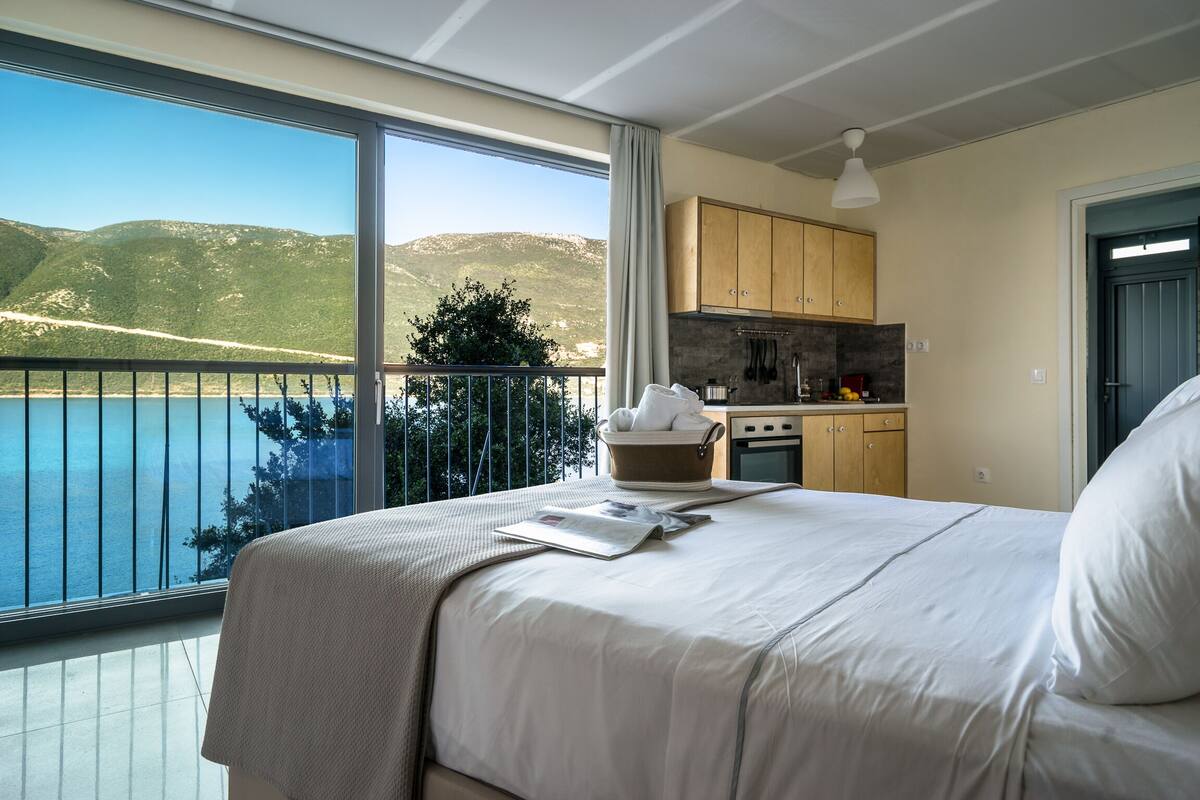 Un dormitorio con un ventanal grande con vistas a un lago y montañas, una kitchenette y un balcón con vistas.