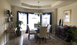 Un coin repas agrémenté d’un lustre, d’une table avec des chaises, d’un buffet et d’une grande fenêtre avec des rideaux.