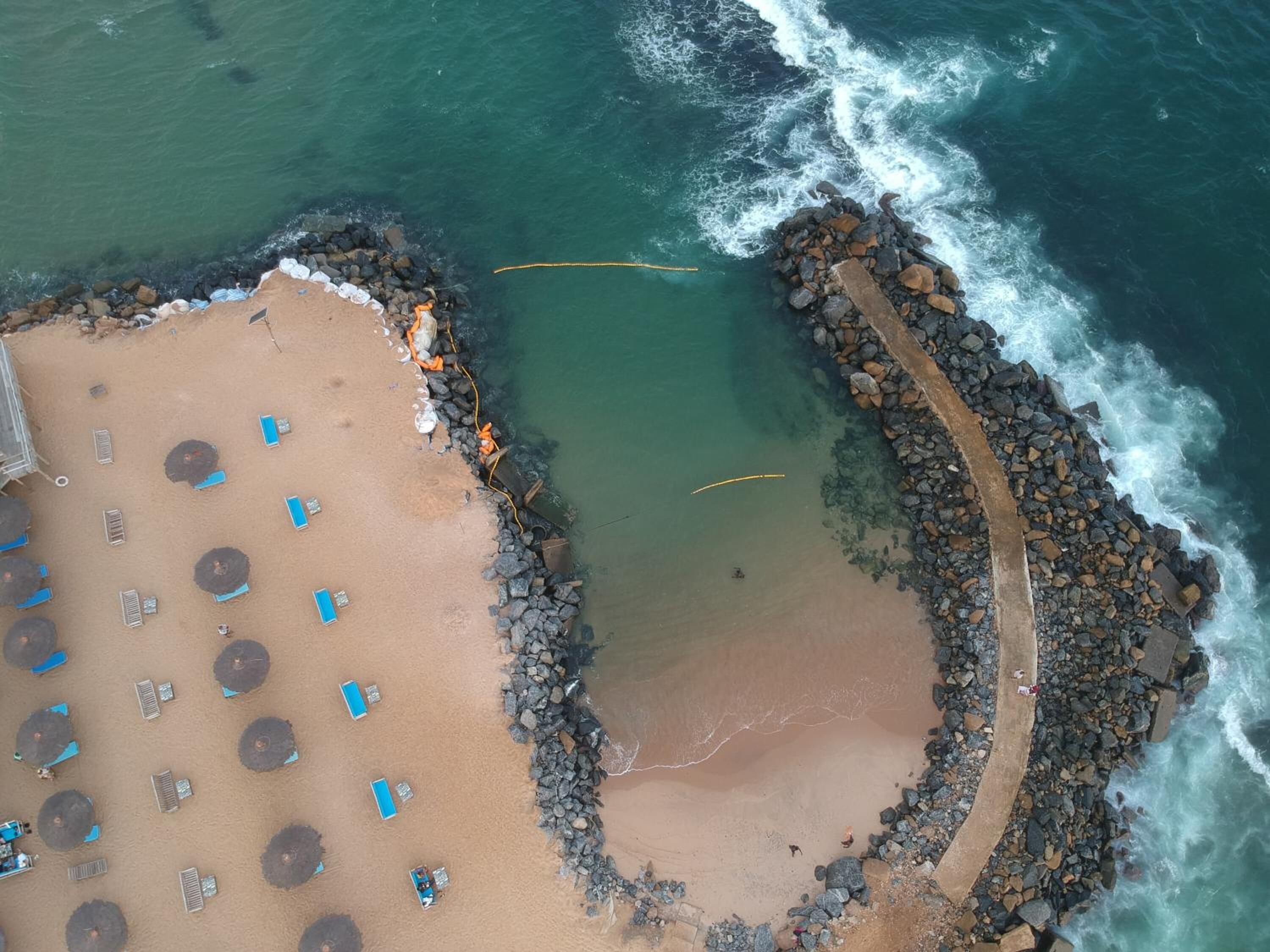 Vista aérea de una playa con tumbonas y sombrillas, un rompeolas rocoso y agua azul cristalina.