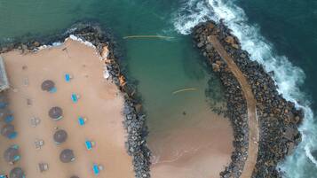 An aerial view of a beach with sun loungers and umbrellas, a rocky breakwater, and clear blue water.