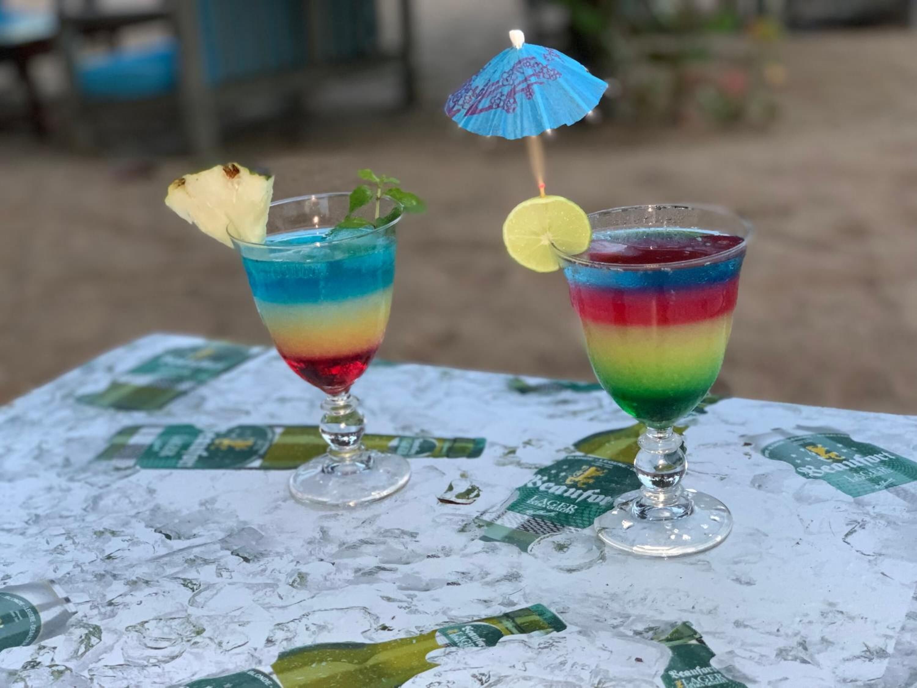 Two colorful drinks on a table with ice and a blue umbrella.