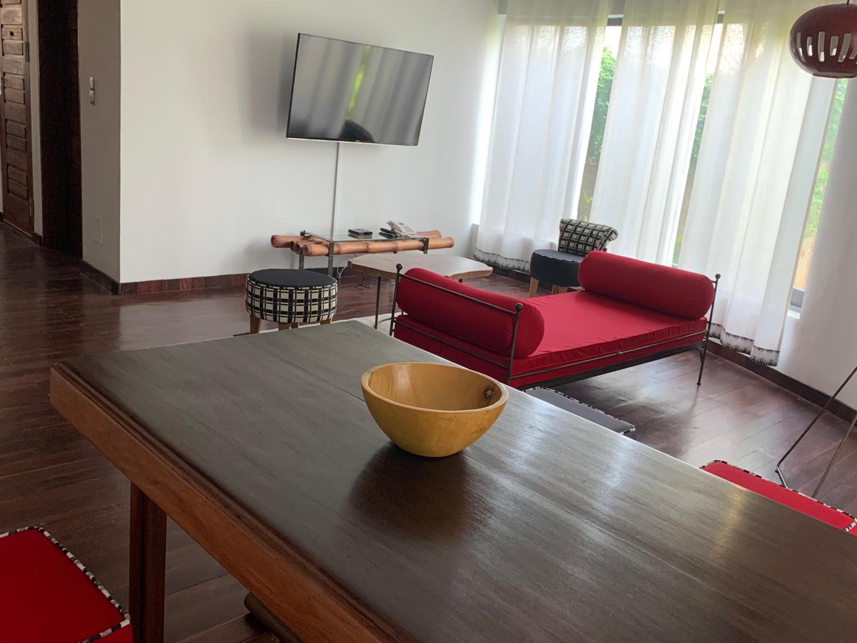 A modern living room with a wooden dining table, a red sofa, a television mounted on the wall, and a window with sheer curtains.