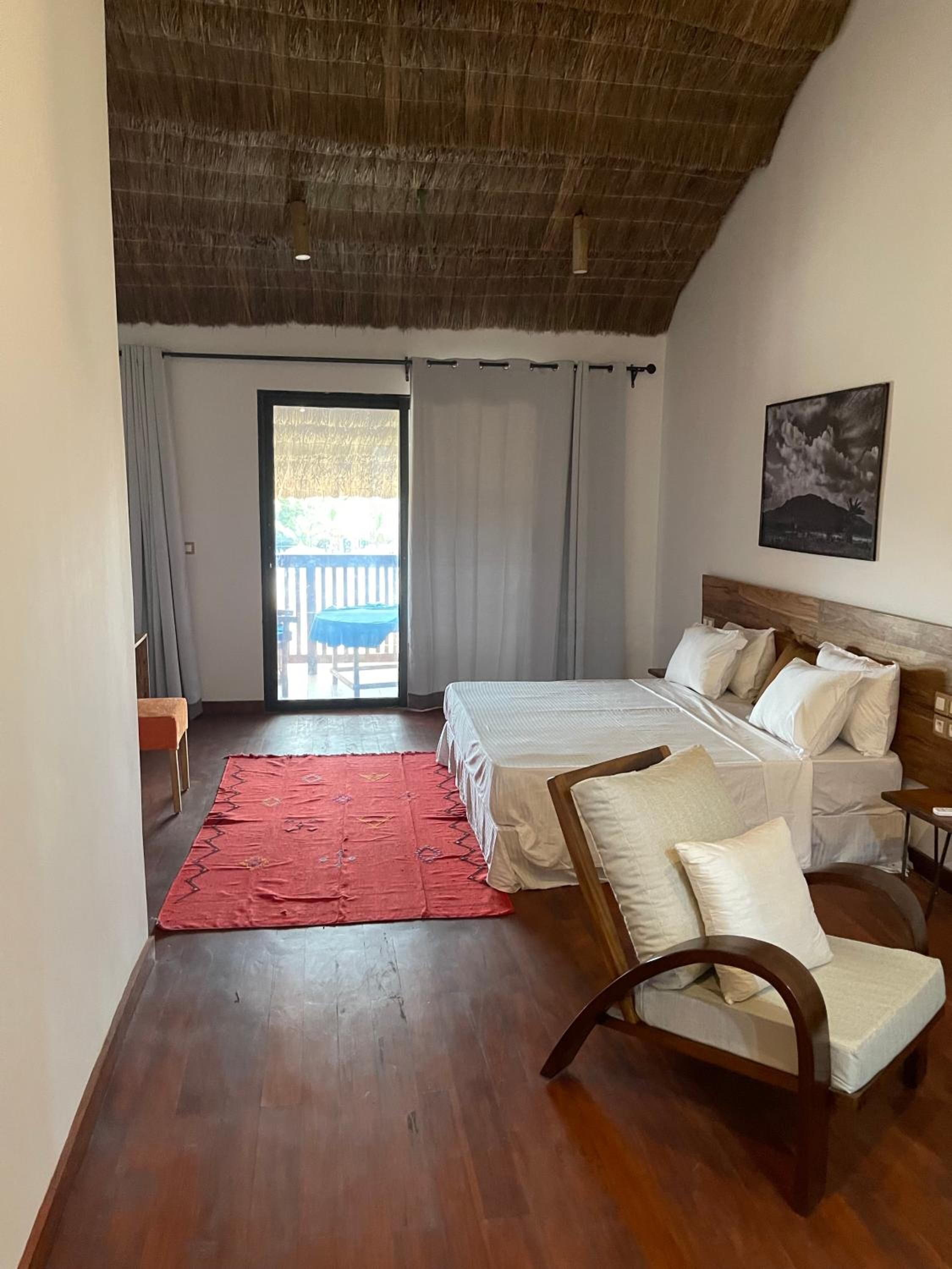 A bedroom with a wooden ceiling, a bed with white bedding, a red rug, a wooden chair, and a view of a balcony with a white fence.