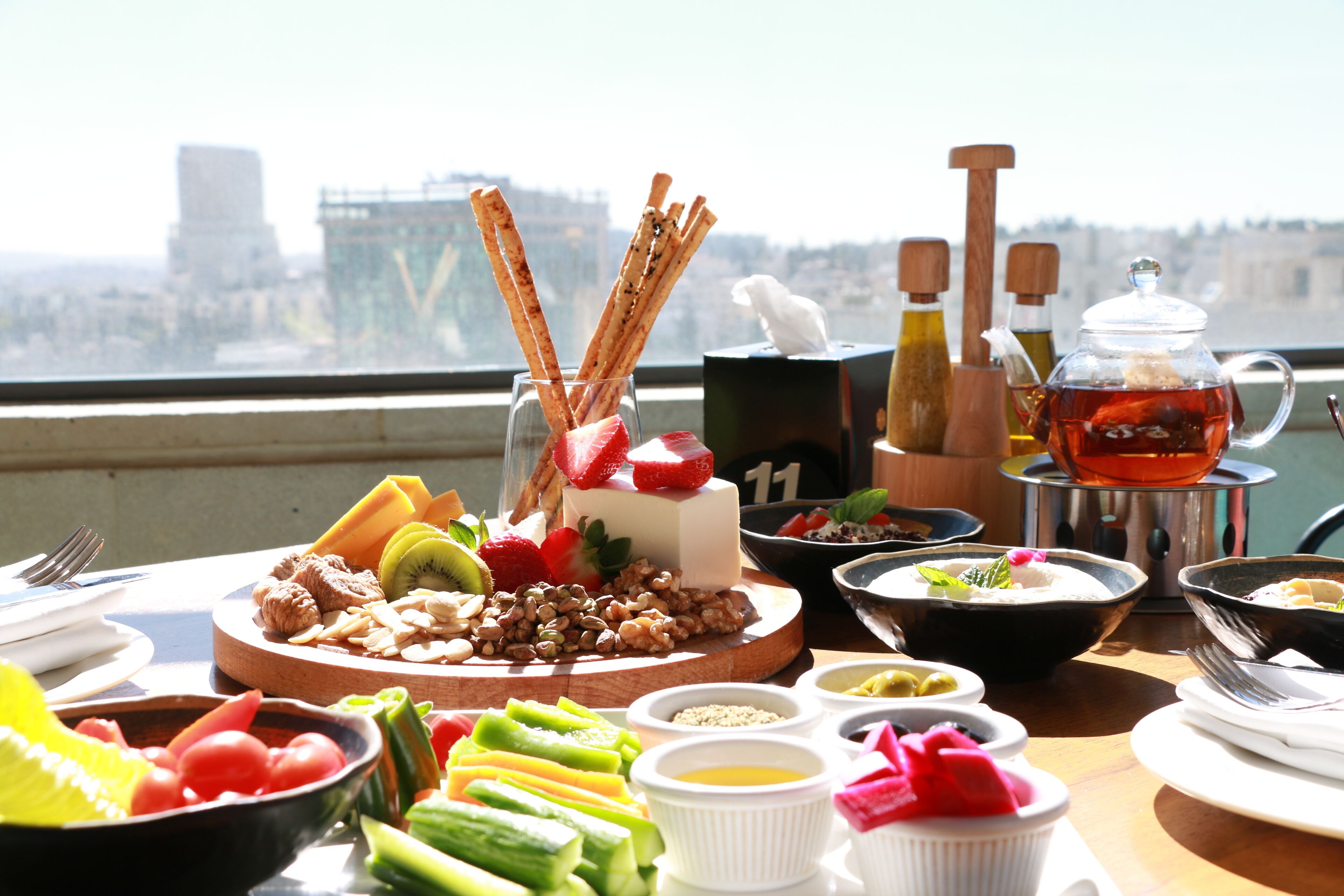 A table with various food items and a teapot, set against a view of a cityscape.