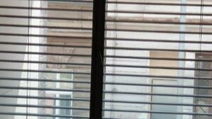 Una ventana con persianas horizontales, un respiradero visible debajo y vistas de un edificio y coches aparcados afuera.