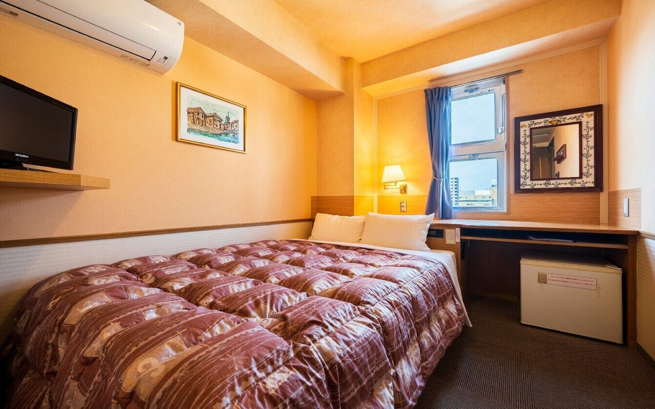 Quarto de hotel com uma cama grande, uma escrivaninha, uma televisão, janela com cortinas e um frigorífico pequeno.