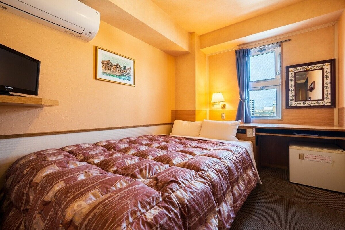 Hotellihuone, jossa on suuri sänky, työpöytä, televisio, ikkuna verhoilla ja pieni jääkaappi.