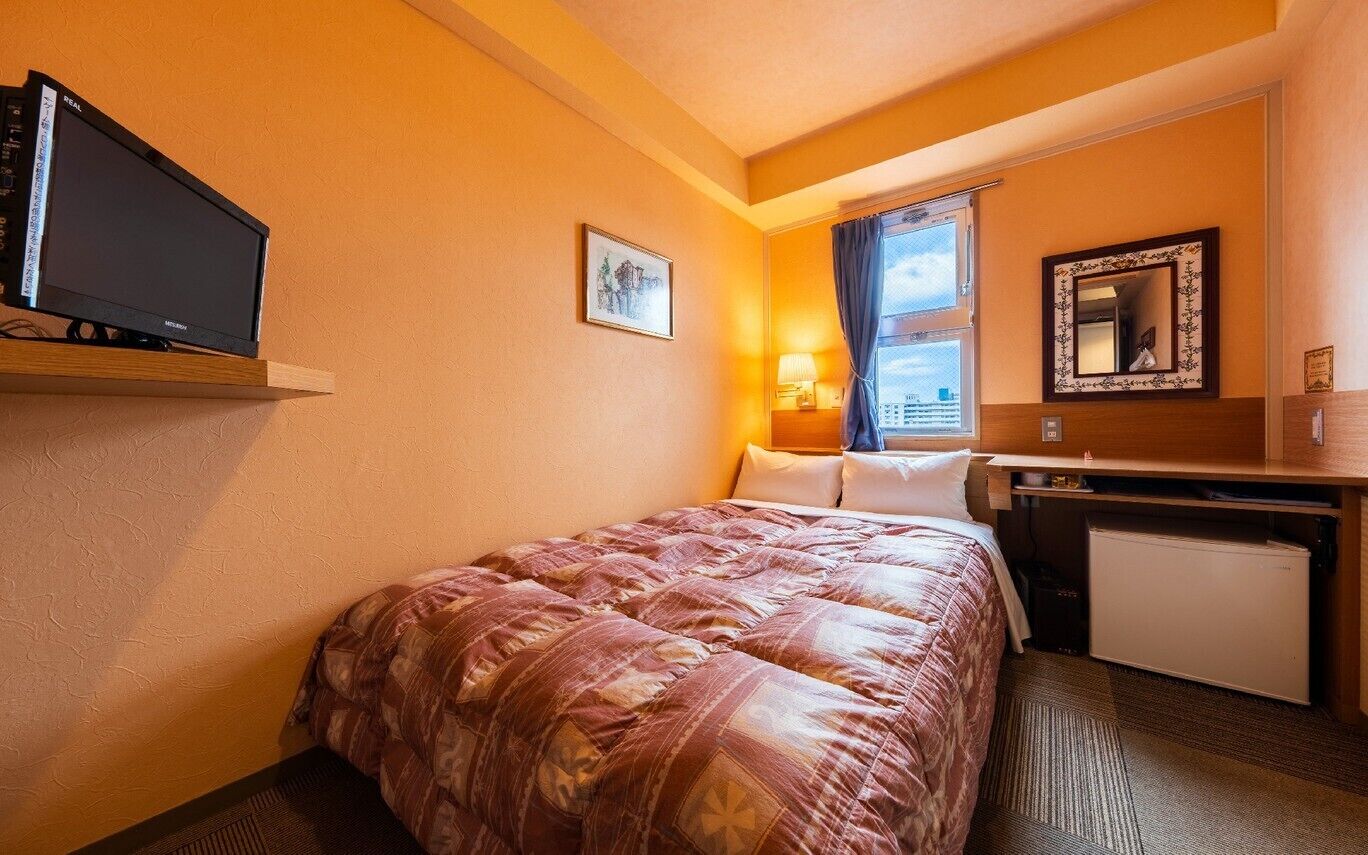 Quarto de hotel com cama, televisão, escrivaninha, espelho e janela com cortinas.