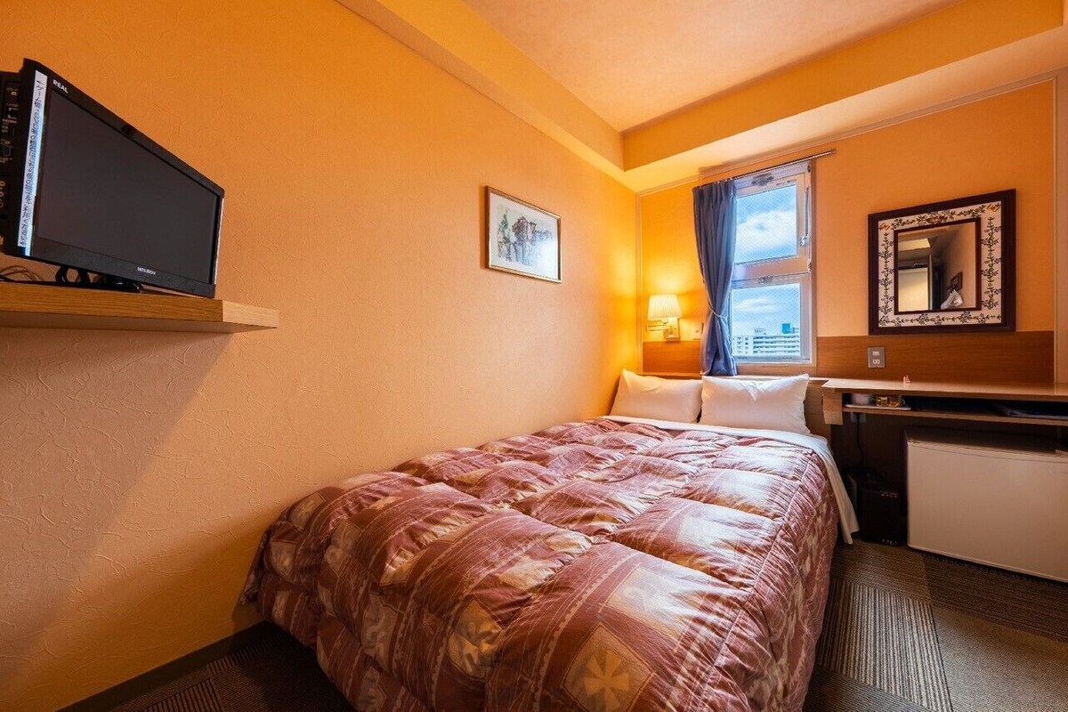 Hotellihuone, jossa on sänky, televisio, työpöytä, peili ja ikkuna, jossa on verhot.