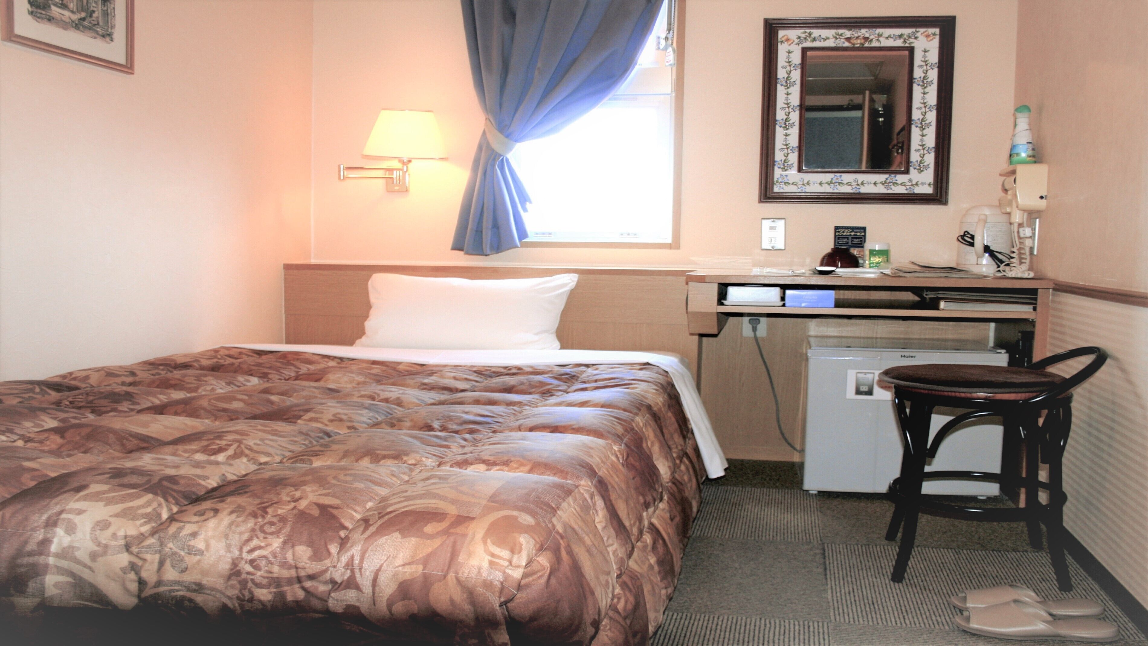 Quarto de hotel com cama, escrivaninha, cadeira, pequeno frigorífico e espelho na parede.