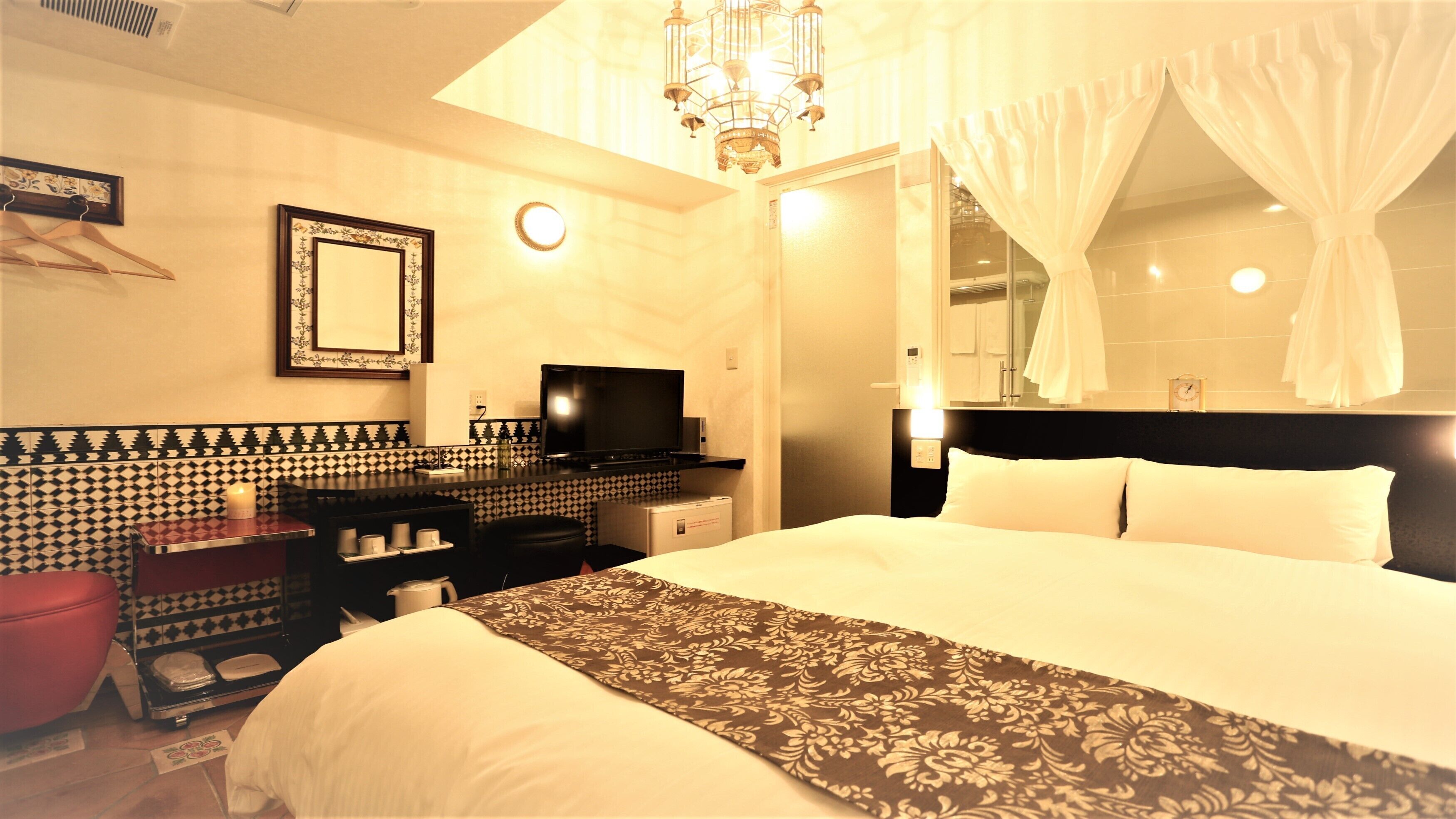 Quarto de hotel com cama, televisão, minibar, espelho e lustre.