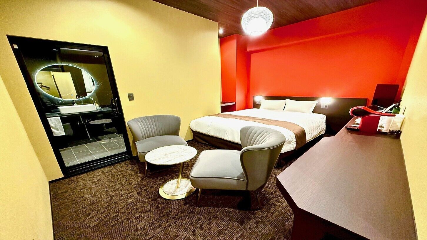 Quarto de hotel com uma cama, duas poltronas, uma mesa redonda pequena, uma escrivaninha, um espelho e uma parede de destaque vermelha.